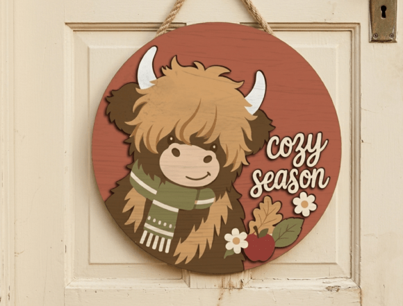 Finished Cozy Season Door Plaque showing a 20cm round wooden sign with a layered Highland cow wearing a winter scarf next to cozy season script lettering.