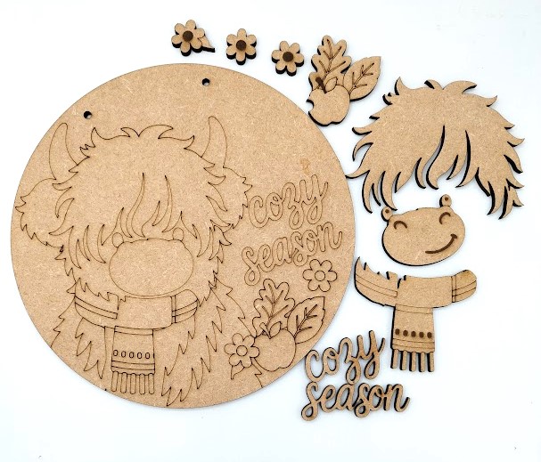 Highland cow wood craft kit Cozy Season Door Plaque showing a 20cm round wooden sign with a layered Highland cow wearing a winter scarf next to cozy season script lettering.
